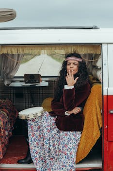 A woman in boho fashion smokes beside a decorated campervan, embracing a retro vibe.