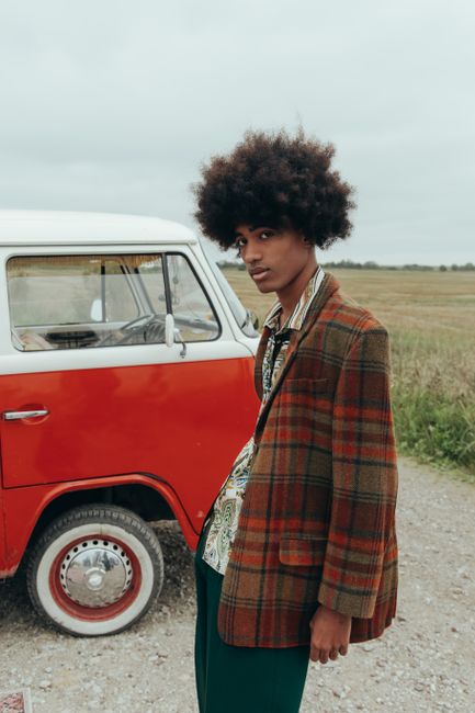 Fotomural Hombre joven con cabello afro en un blazer retro posando junto a una furgoneta roja clásica al aire libre.