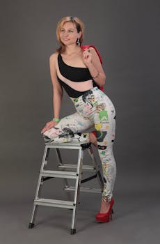 Stylish woman in unique clothing posing on a stepladder in a studio environment.