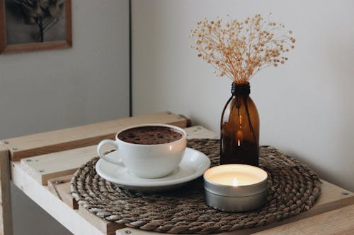 A Cup of Coffee Near the Glass Bottle with Dried Flowers and Lighted Candle on a Woven Mat