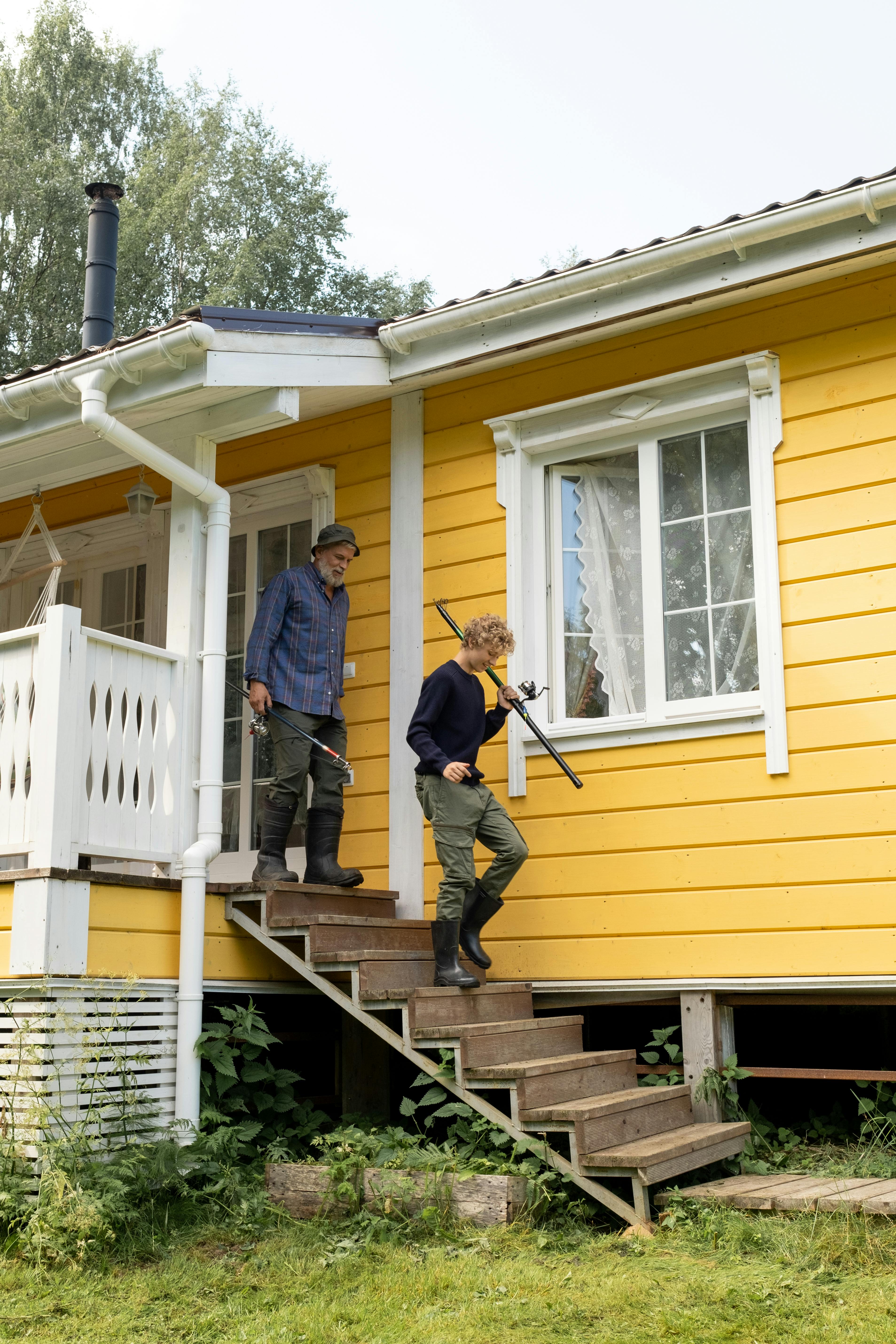 Grandfather and grandson with fishing rods departing cozy yellow cottage for outdoor adventure.