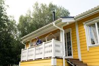 A senior man enjoys a peaceful afternoon on a sunlit yellow house balcony surrounded by trees.