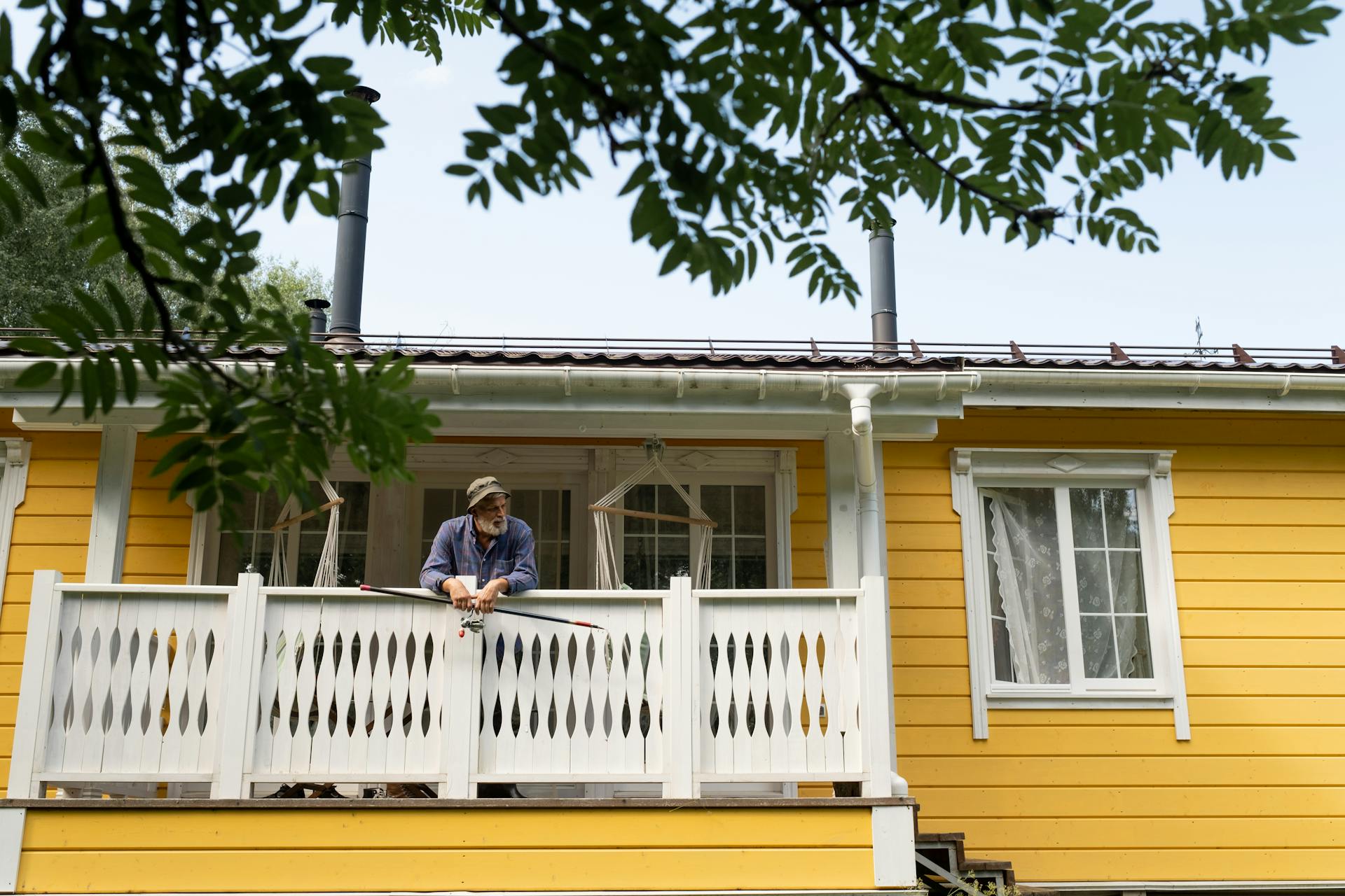 A senior man enjoys a quiet moment on a yellow country house balcony with a fishing rod.