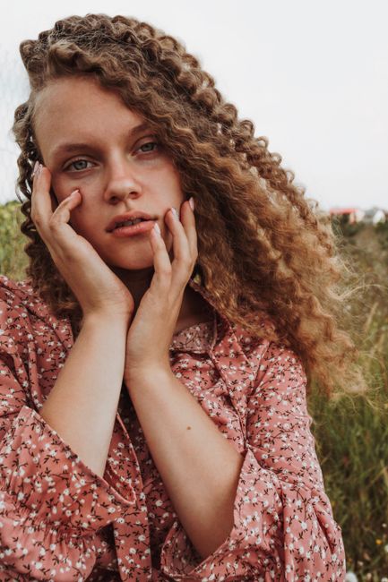 Portrait of a young woman with curly hair posing in a floral dress, outdoors in a natural setting.