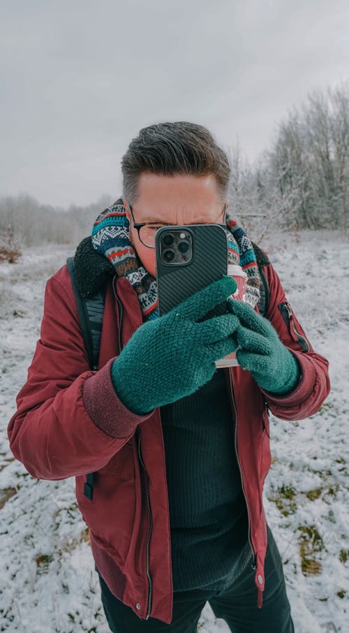 Free A Man in Red Jacket Holding His Mobile Phone while Standing on a Snow Covered Ground Stock Photo