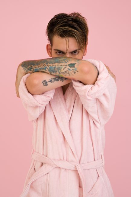A modern and stylish portrait of a tattooed model posing in a pink bathrobe against a matching background.