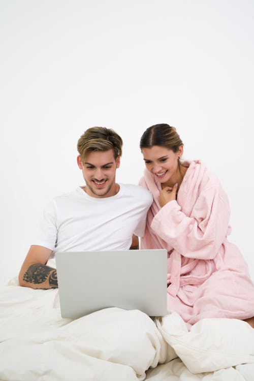 Man in White Crew-neck Shirt Sitting on Bed Beside Woman in Pink Bathrobe Looking at Laptop Computer