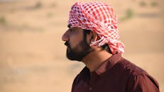 一名戴着传统红白头巾、留着浓密胡须的男子站在沙漠中，侧面轮廓捕捉到了他的形象