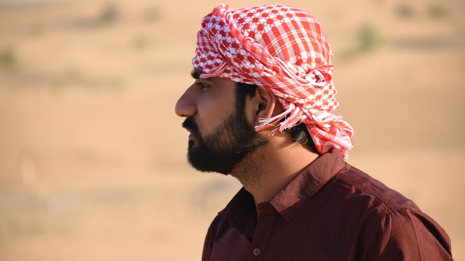A man wearing a traditional red and white headscarf stands in the desert with a full beard, captured in a side profile.