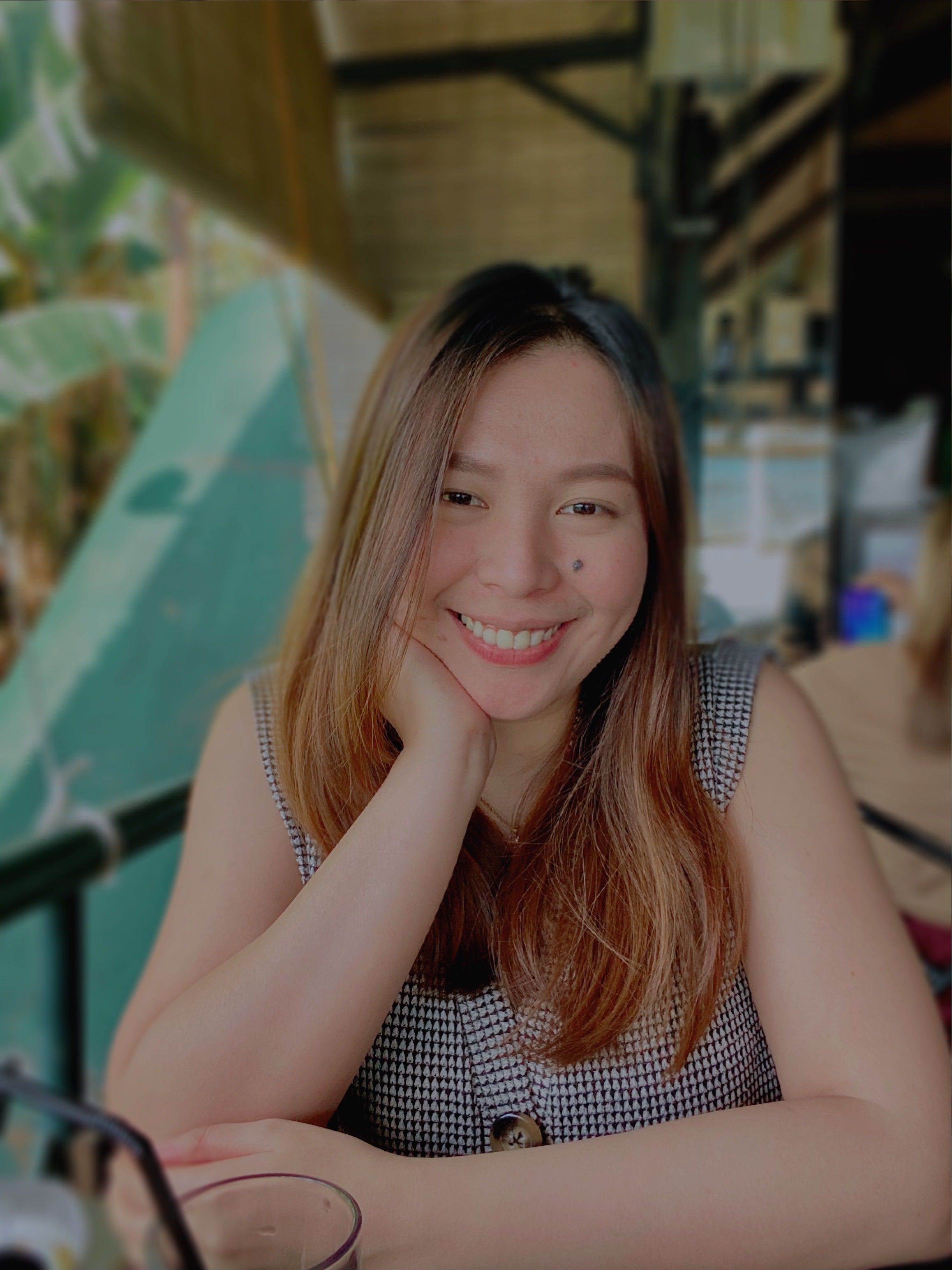 Portrait of a smiling woman in a sleeveless shirt, seated at a café with a tropical backdrop.