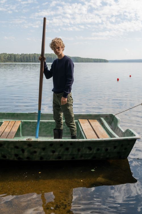 Free A young boy stands with an oar on a small boat in a calm lake, under a partly cloudy sky. Stock Photo