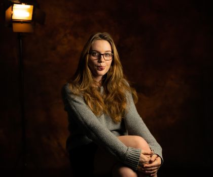 Lighthearted studio photo of a woman sitting with glasses and a playful expression.