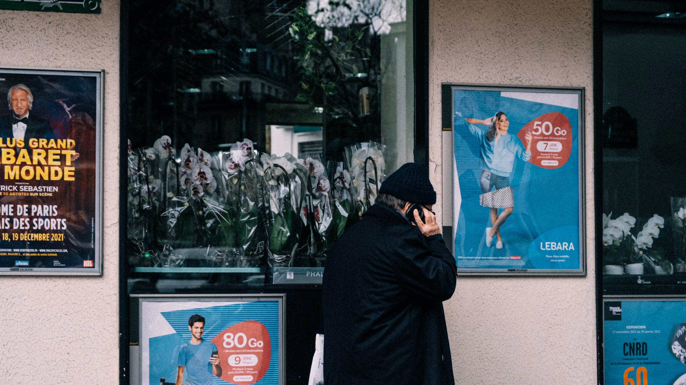 An adult male making a phone call outside a shop with posters and reflections.