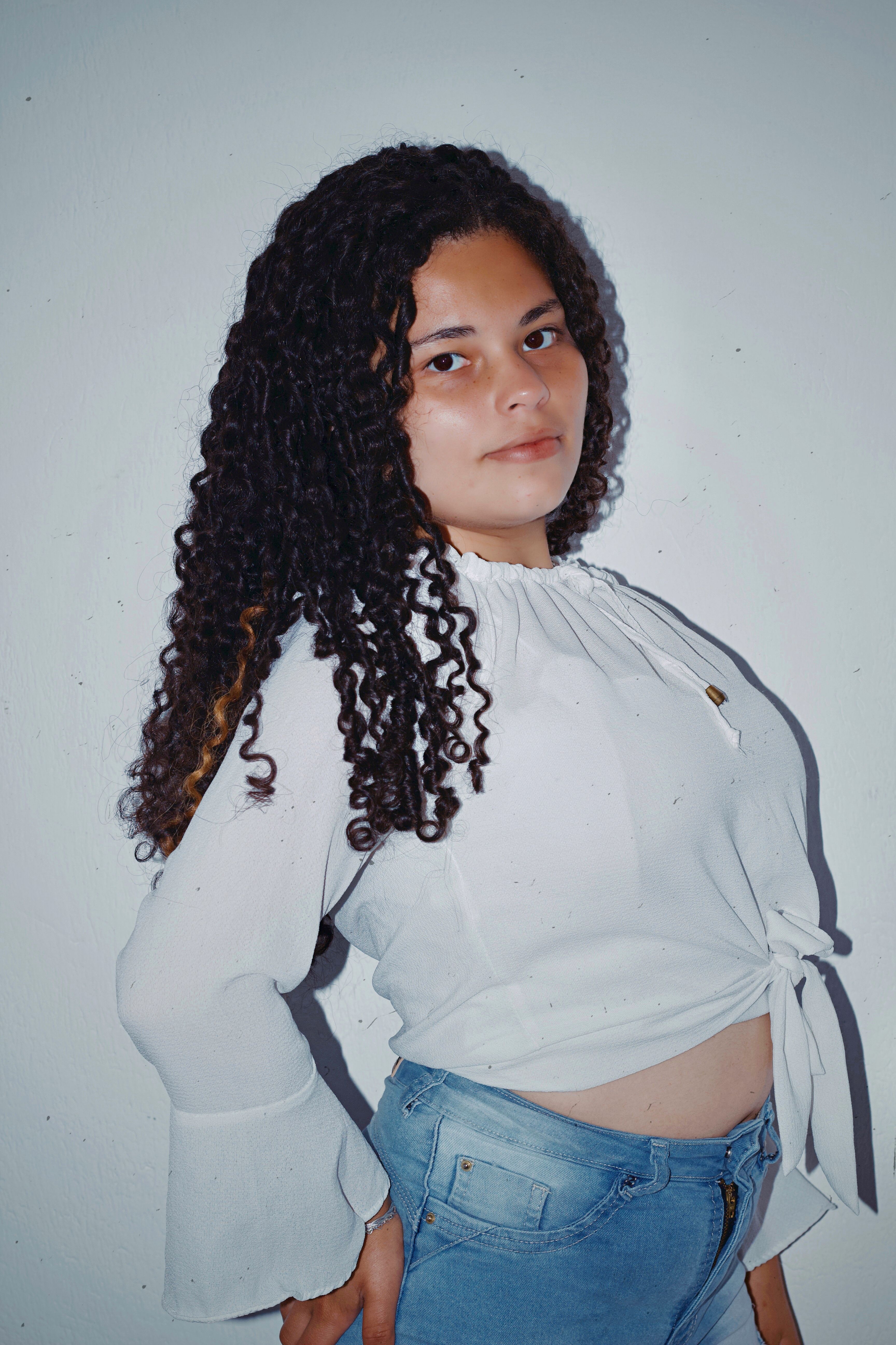 A confident woman with curly hair poses in a stylish white blouse and jeans.