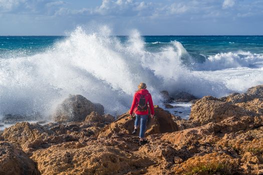 A woman stands on a rocky coast as waves crash, exploring the seashore outdoors.