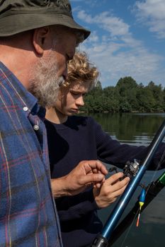 An elderly man teaches his teenage grandson how to fish on a sunny day by the lake.