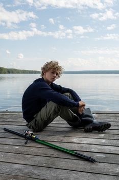 Blond teenager sitting on a pier, with curly hair, fishing rod, and boots, enjoying a calm day by the lake.