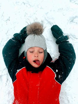 Young child in winter attire playfully sticks tongue out while lying in snow.