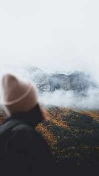 A person in a beanie looks at a snow-covered mountain surrounded by clouds and autumn foliage.