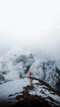 A lone hiker in an orange jacket stands on a snowy mountain ridge under a cloudy sky.