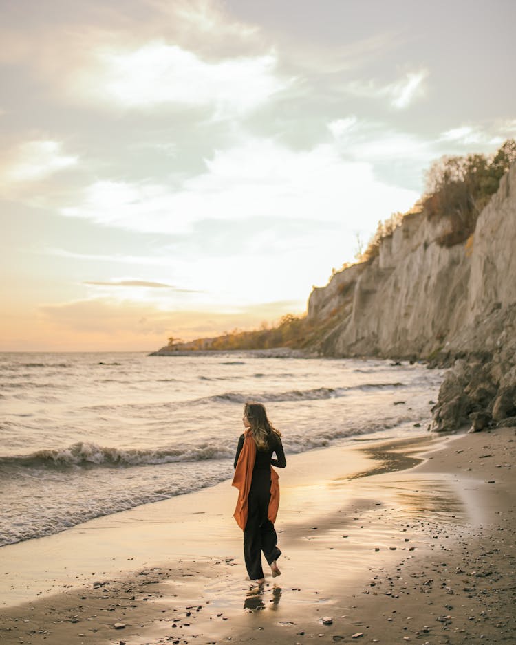 Woman In Scarf Walking On Beach Towards Cliff At Sunset