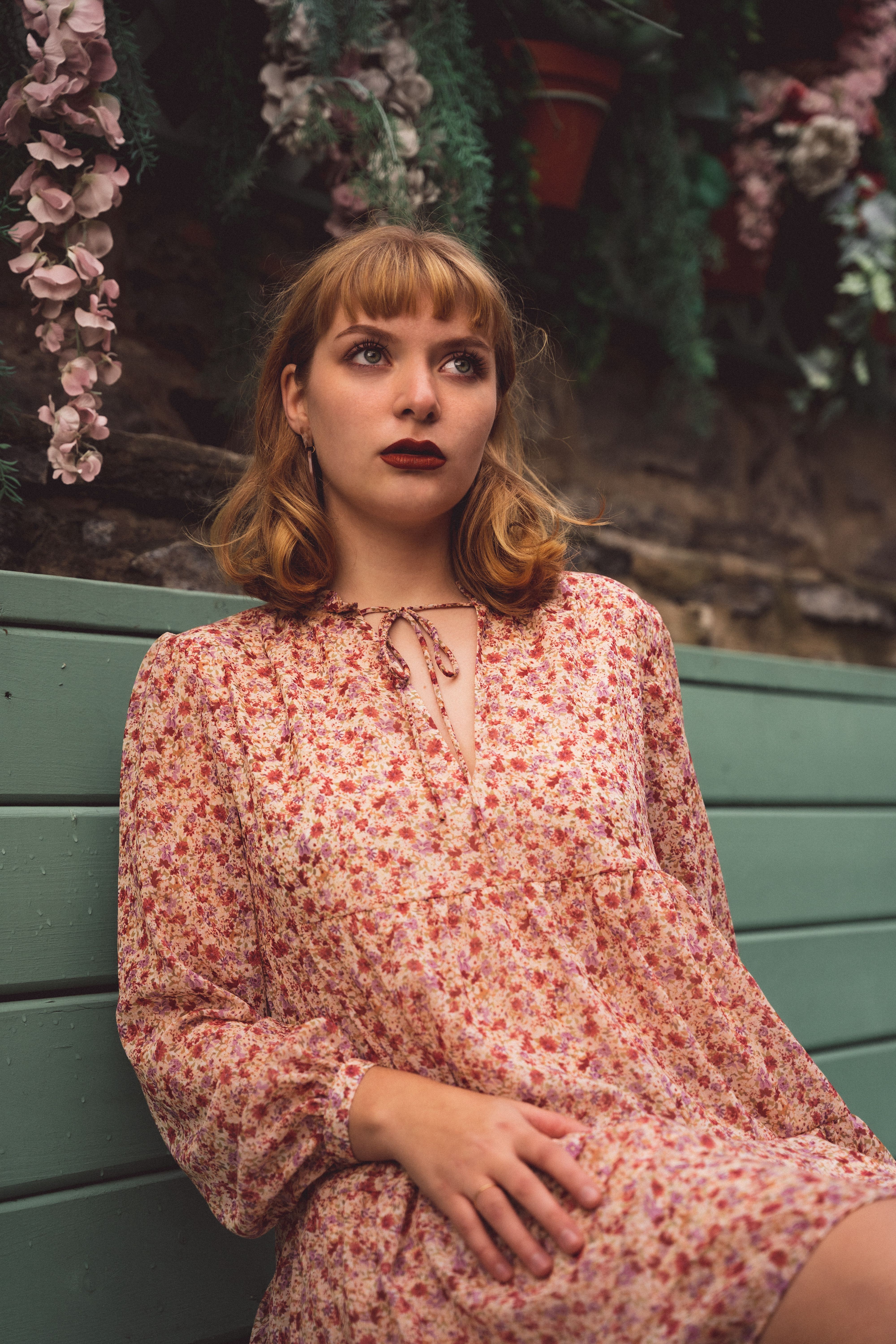 A stylish woman in a floral dress sits on a bench surrounded by greenery.