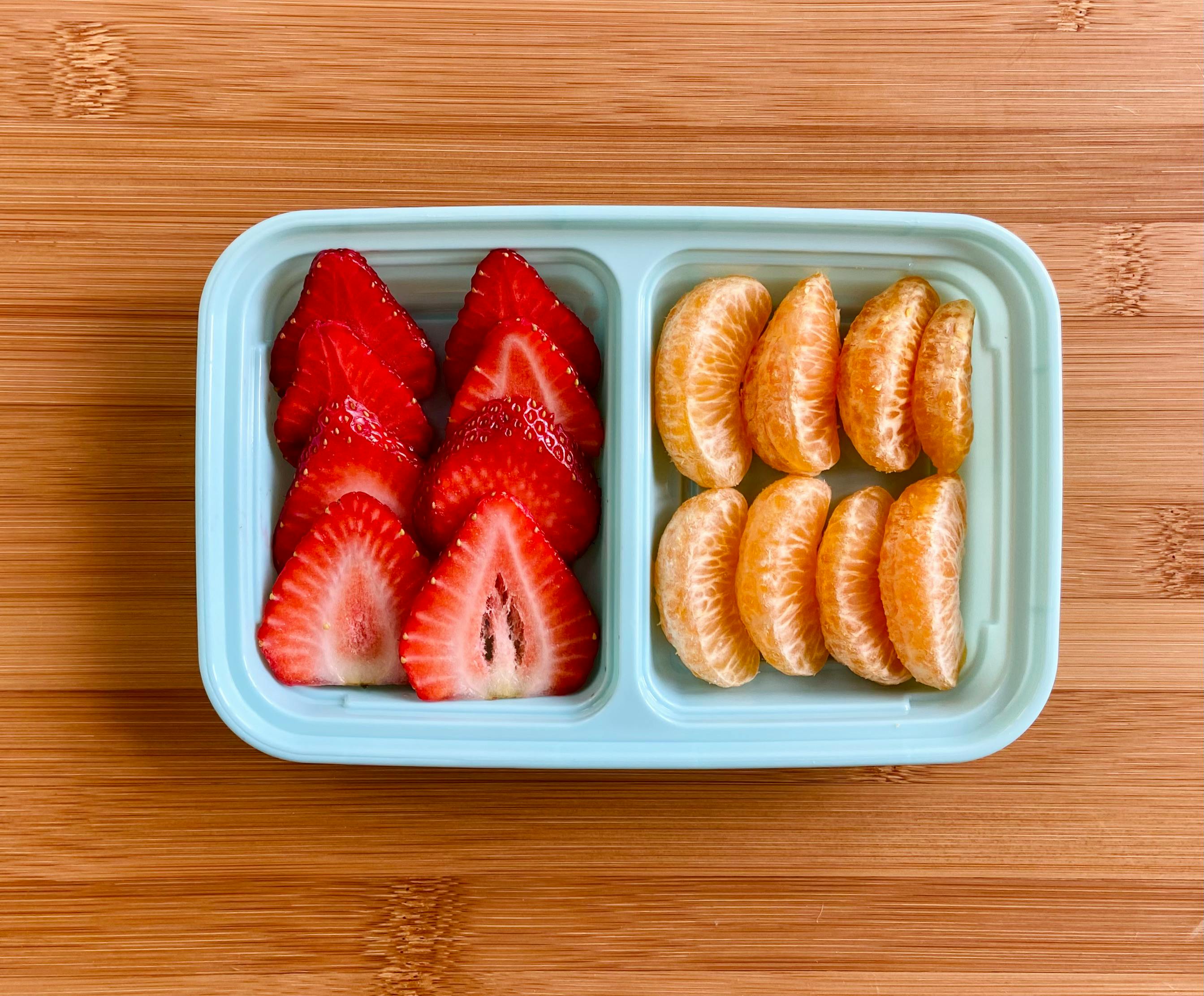 A healthy snack of sliced strawberries and orange segments in a bento box on a wooden background.