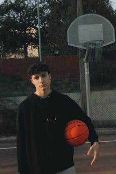 Teenage boy holding a basketball on an outdoor court at dusk. Casual sports scene.