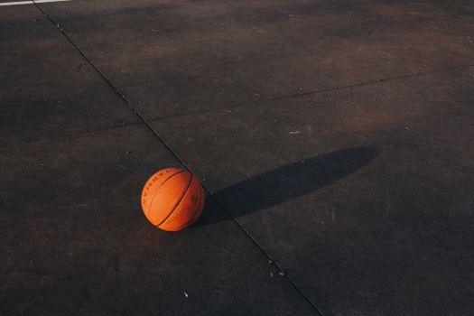 A single basketball casting a shadow on a textured outdoor court surface at dusk.