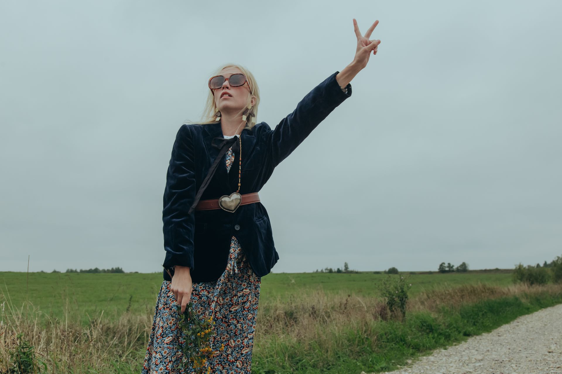 Woman in 70s hippie fashion posing with peace sign in open field.