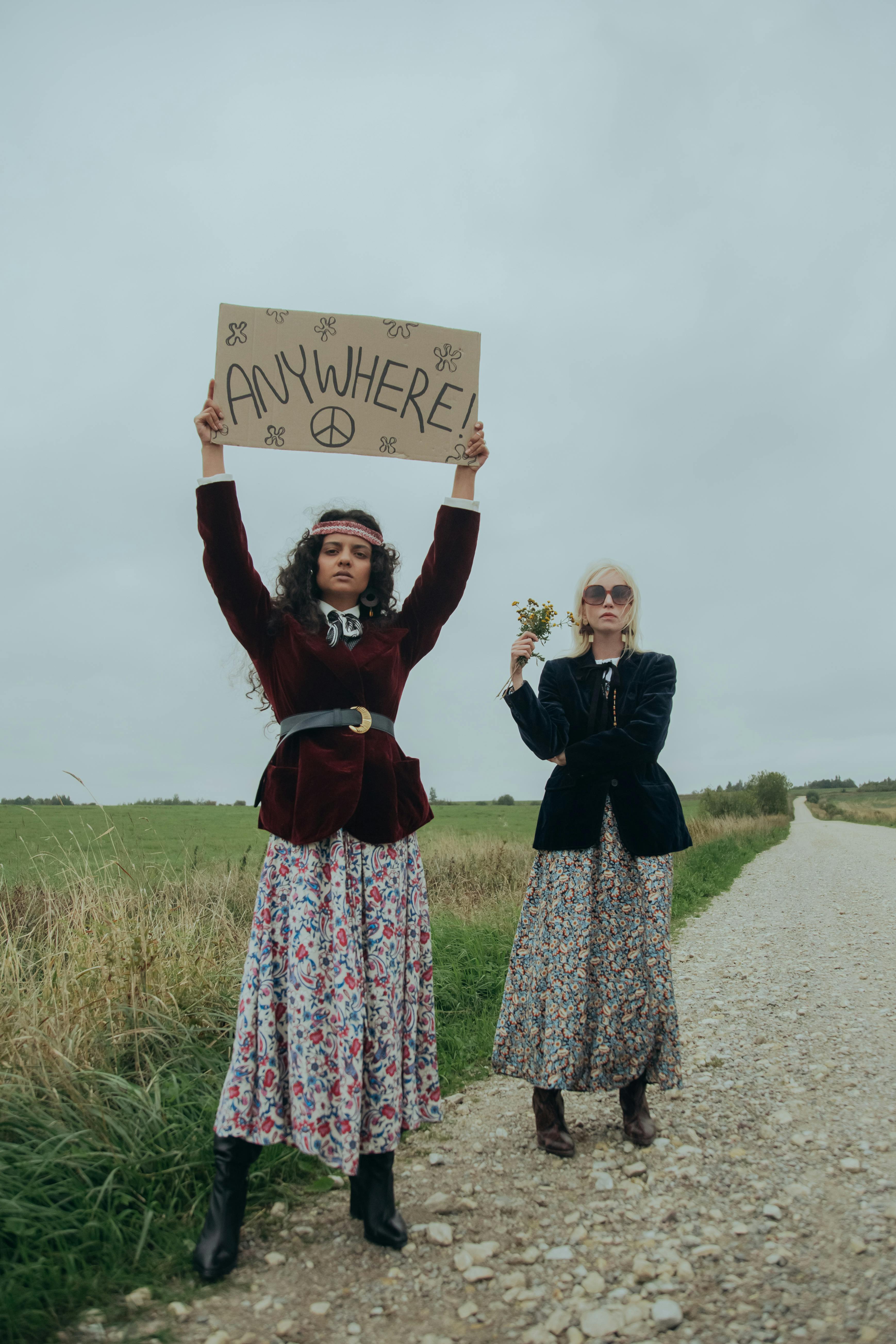 Two women in vintage hippie outfits hitchhiking on a rural road, evoking nostalgia and freedom.