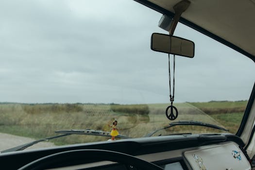 A vintage car interior with a peace symbol decor and a doll on the dashboard, set in a rural landscape.