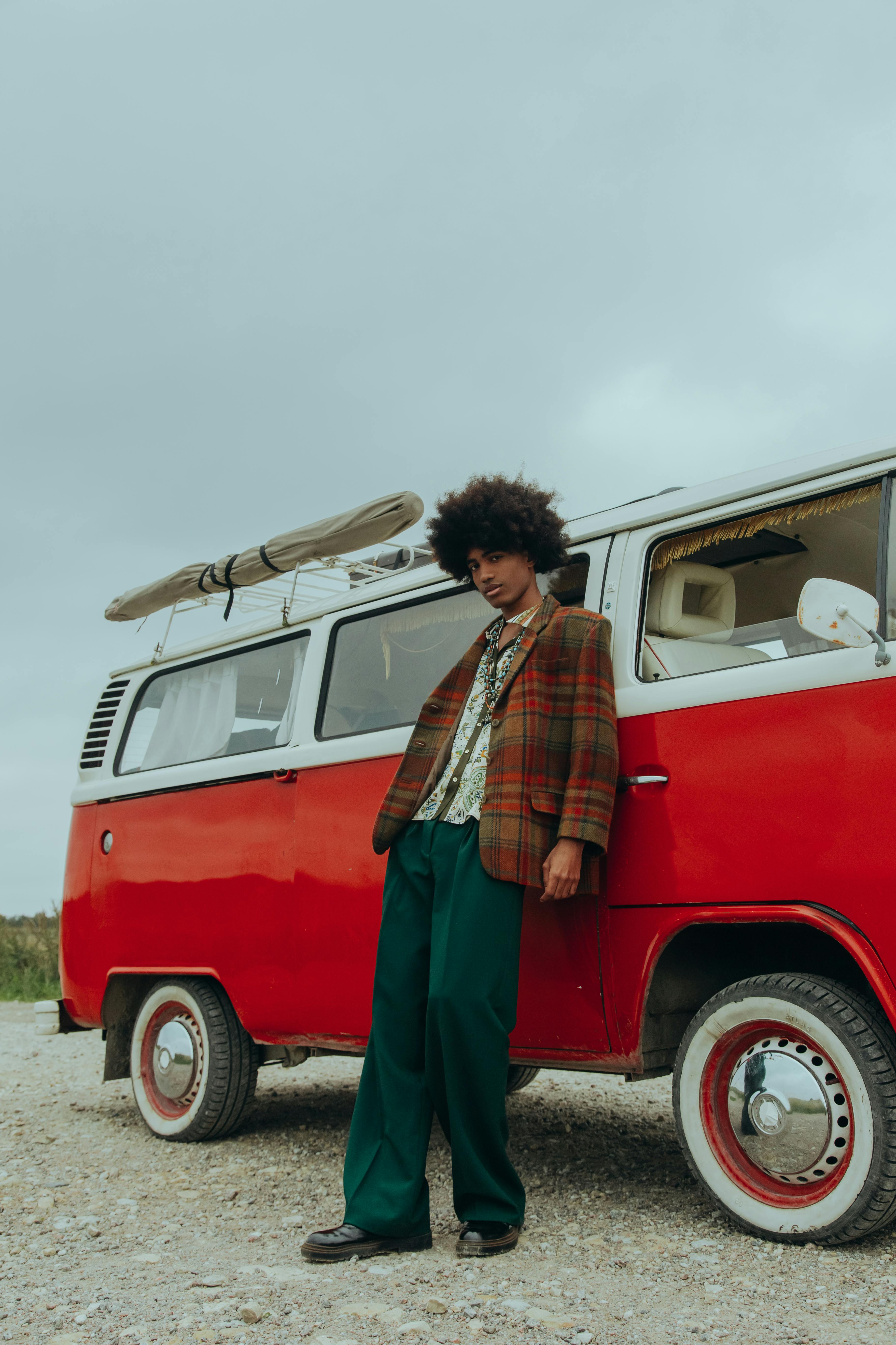 Stylish man with afro leaning on a retro red van outdoors under a cloudy sky.