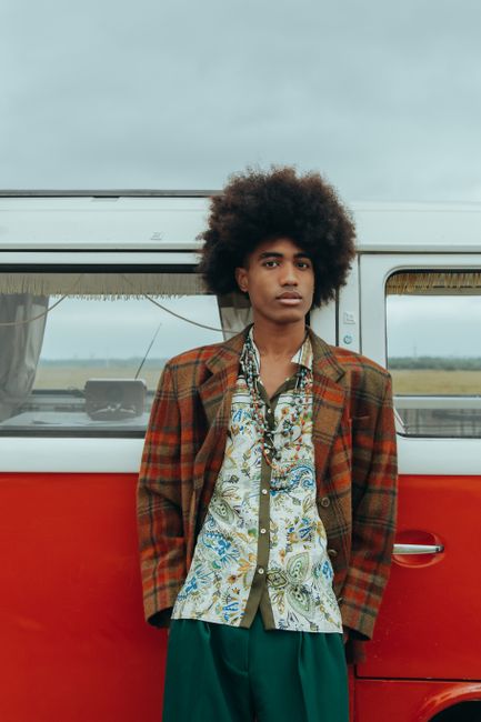 A fashionable afro-haired man in vintage attire leaning against a classic van outdoors.