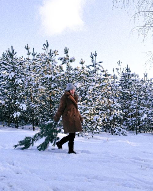 Free A woman in a winter coat and beanie walks across a snow-covered field carrying tree branches. Stock Photo