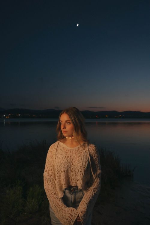 Free A serene portrait of a woman by a lakeside under the crescent moon at twilight. Stock Photo