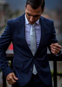 A stylish man in a dark suit looks down, showcasing elegance and sophistication, set against a blurred outdoor background.