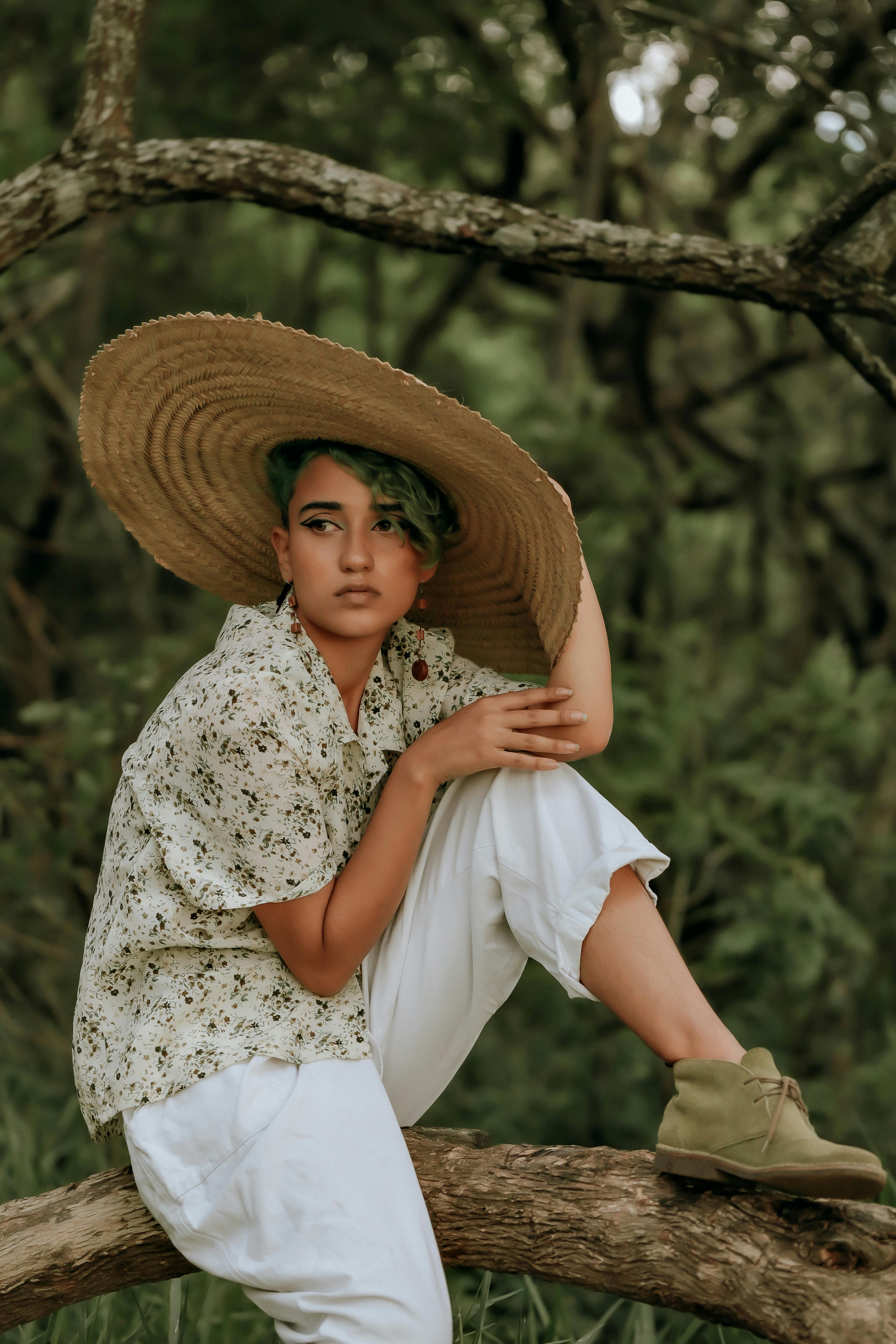 A stylish woman in a floral shirt and large hat sitting outdoors on a tree trunk.