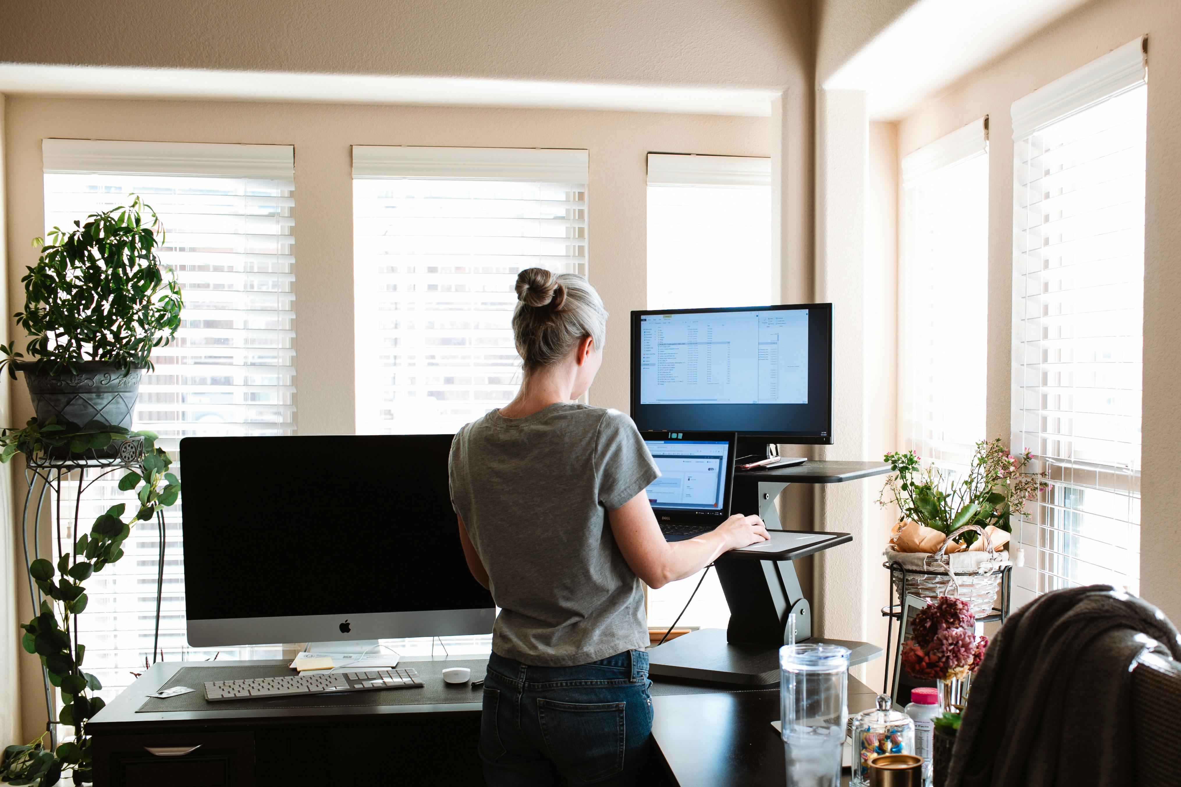 Free A woman standing while using a laptop and monitor in a bright, modern home office space. Stock Photo