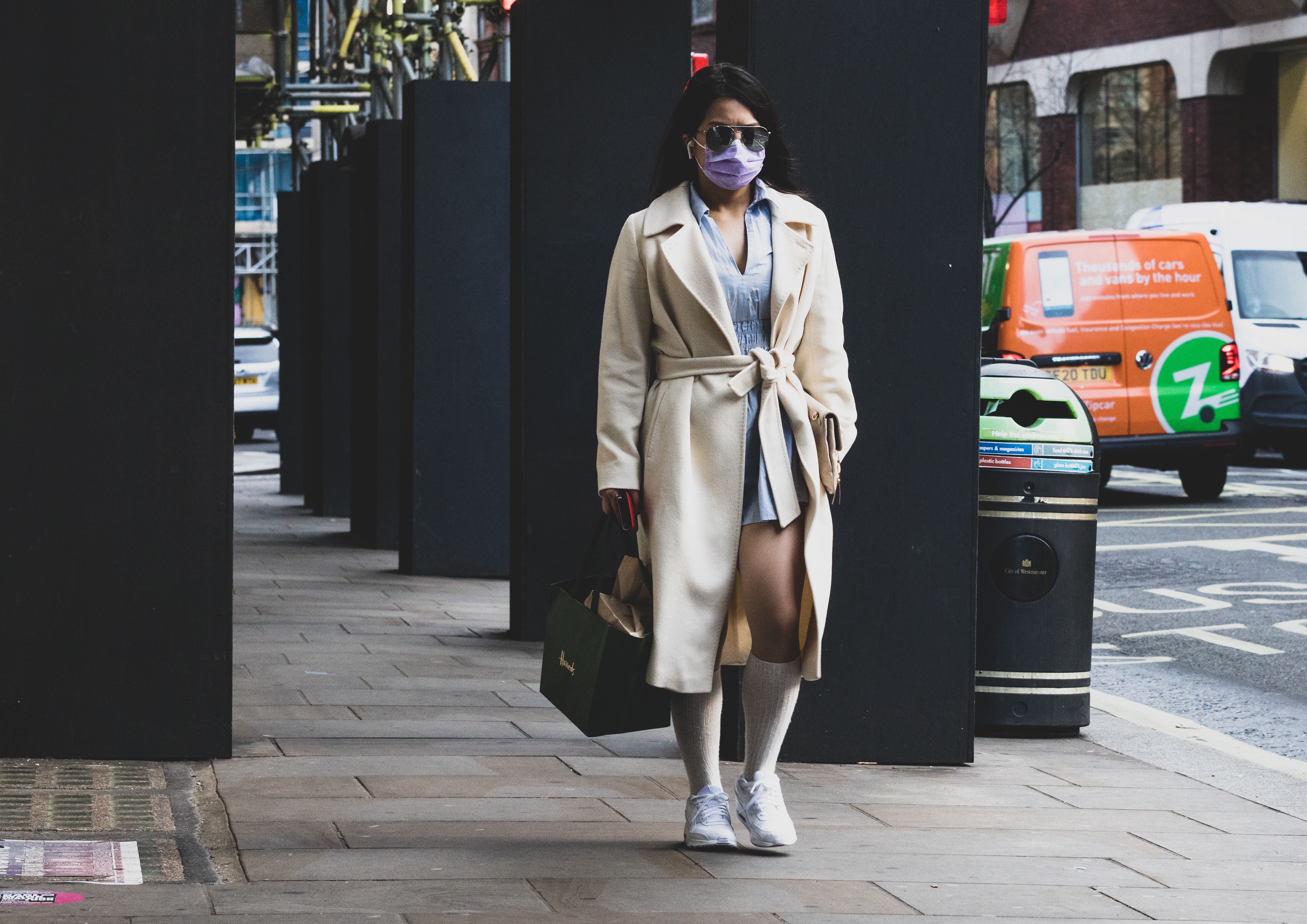 A woman in a coat and face mask walks on an urban street, showcasing fashion and the new normal.