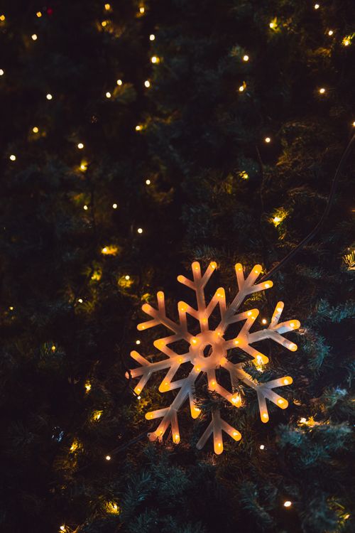 Free Illuminated snowflake ornament on Christmas tree with string lights creating a cozy winter atmosphere. Stock Photo