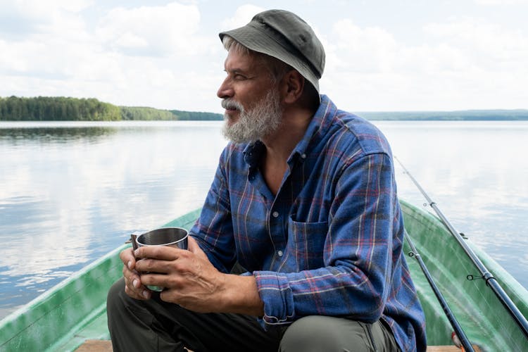 Bearded Senior Man In Hat Sitting In Boat And Holding Mug 