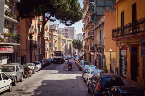 Free A sunlit urban street lined with parked cars and colorful buildings, perfect for city exploration. Stock Photo