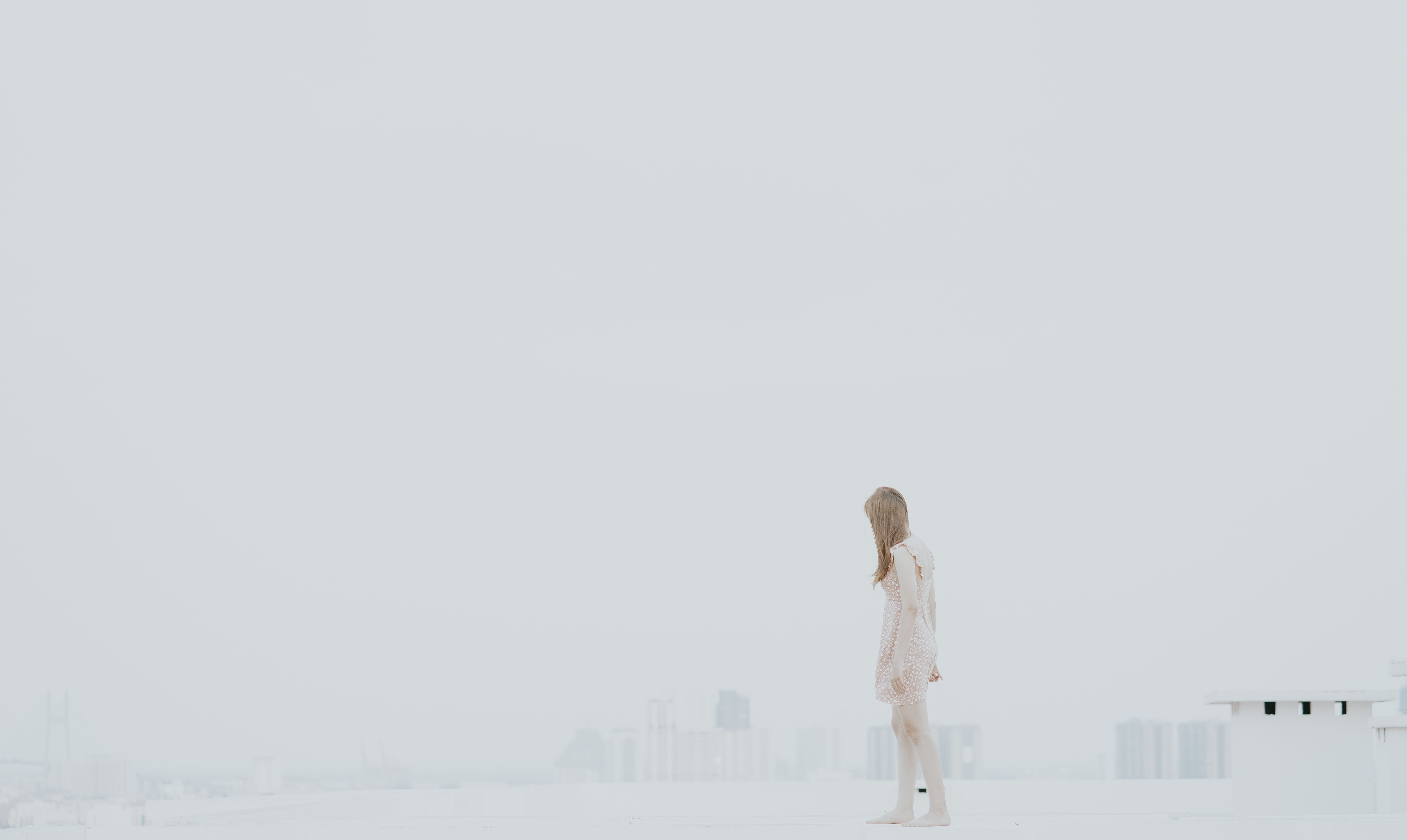 A woman in a beige dress stands barefoot on a rooftop with a foggy cityscape in the background.