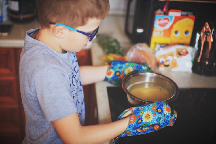 Boy In Glasses And Oven Gloves Carrying A Bowl Of Soup