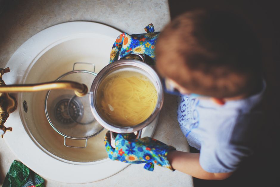 A child strains spaghetti at the sink wearing colorful oven mitts, highlighting a child's culinary exploration.