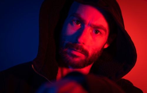 Free Portrait of a man wearing a hood with dynamic neon lighting creating a moody effect. Stock Photo