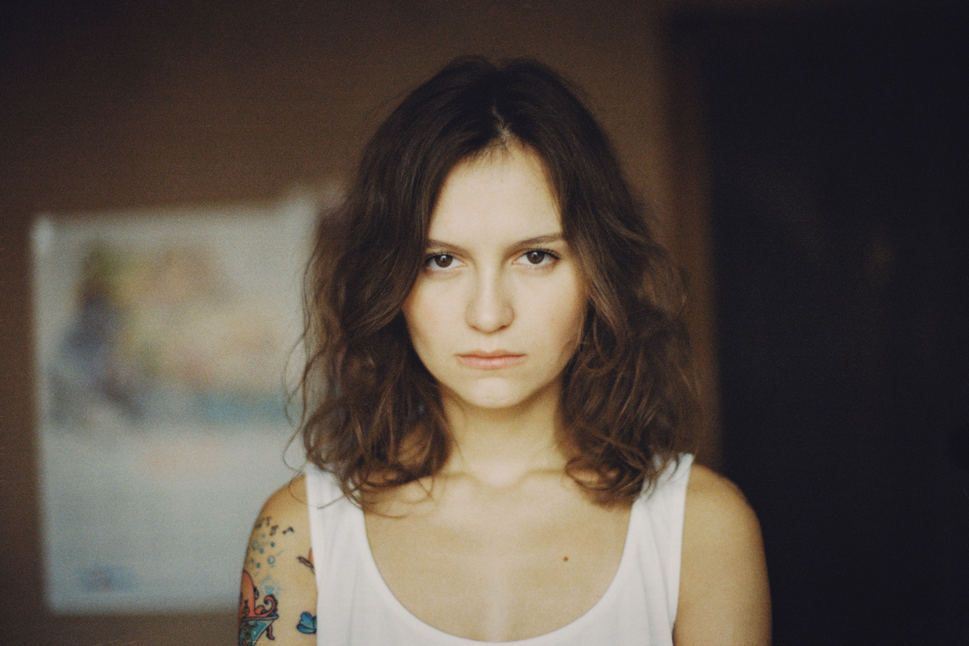 Close-up portrait of a serious woman with arm tattoo in a white top, evoking introspection.