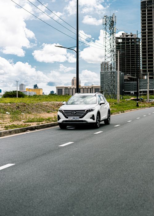 Free White SUV driving on a city road under a blue sky with clouds and skyscrapers in view. Stock Photo