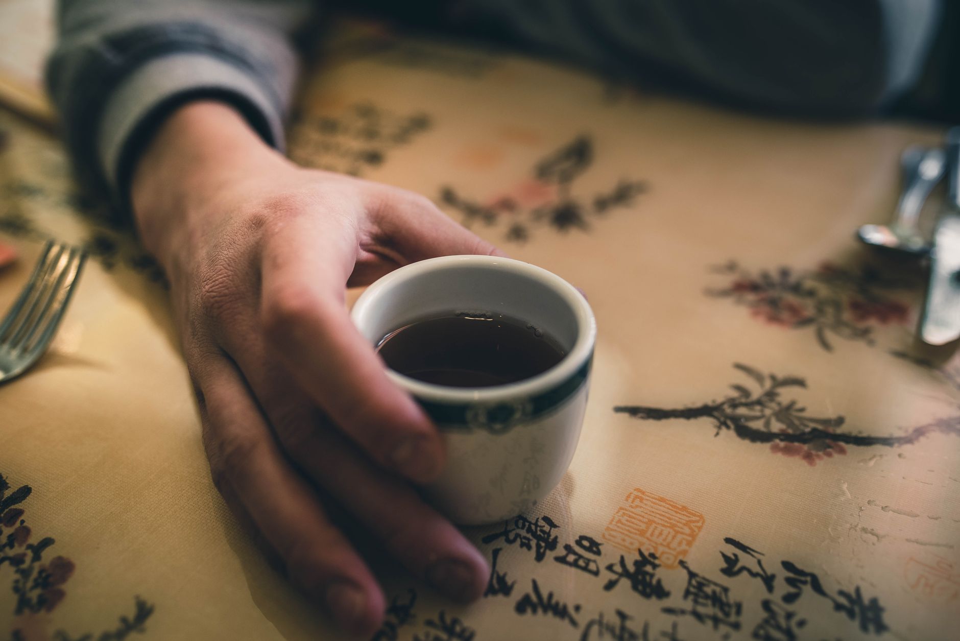 A close-up of a hand holding a cup of hot tea on a table with artistic Asian prints.
